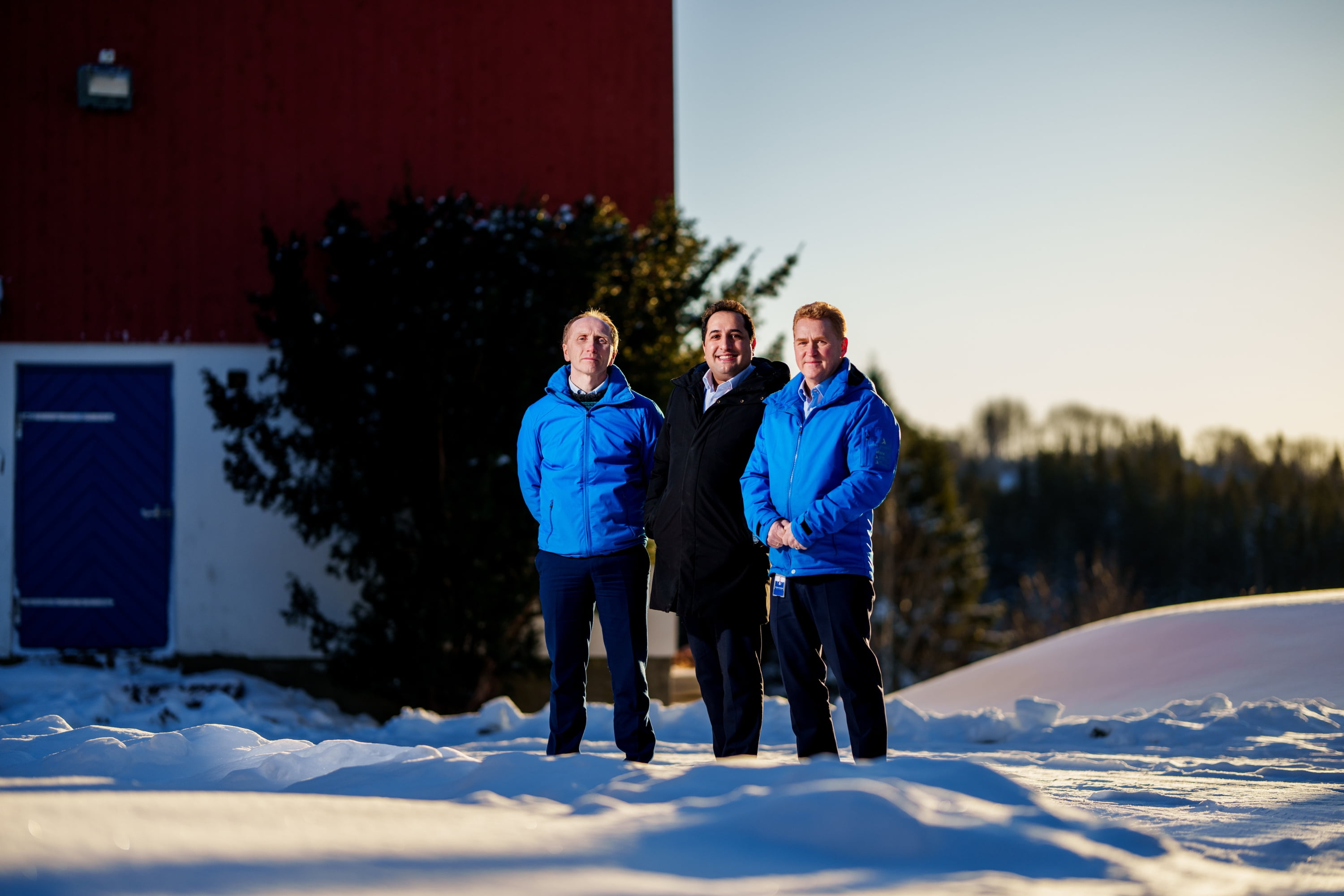 Tre menn i jobbantrekk står foran et rødt fjøs på en vinterdag