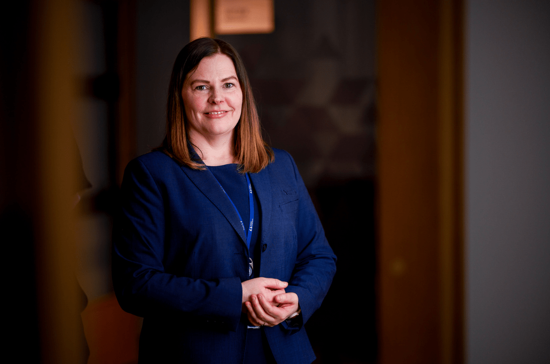 Kvinne står smilende i en gang med hendene i fold.